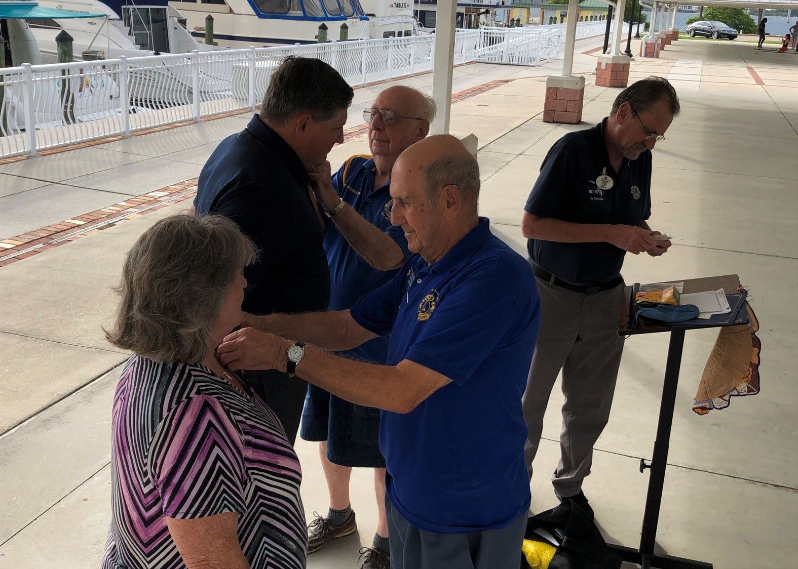 Five people are gathered outside by boats. Two men attach a pin to a woman's collar while another man watches.