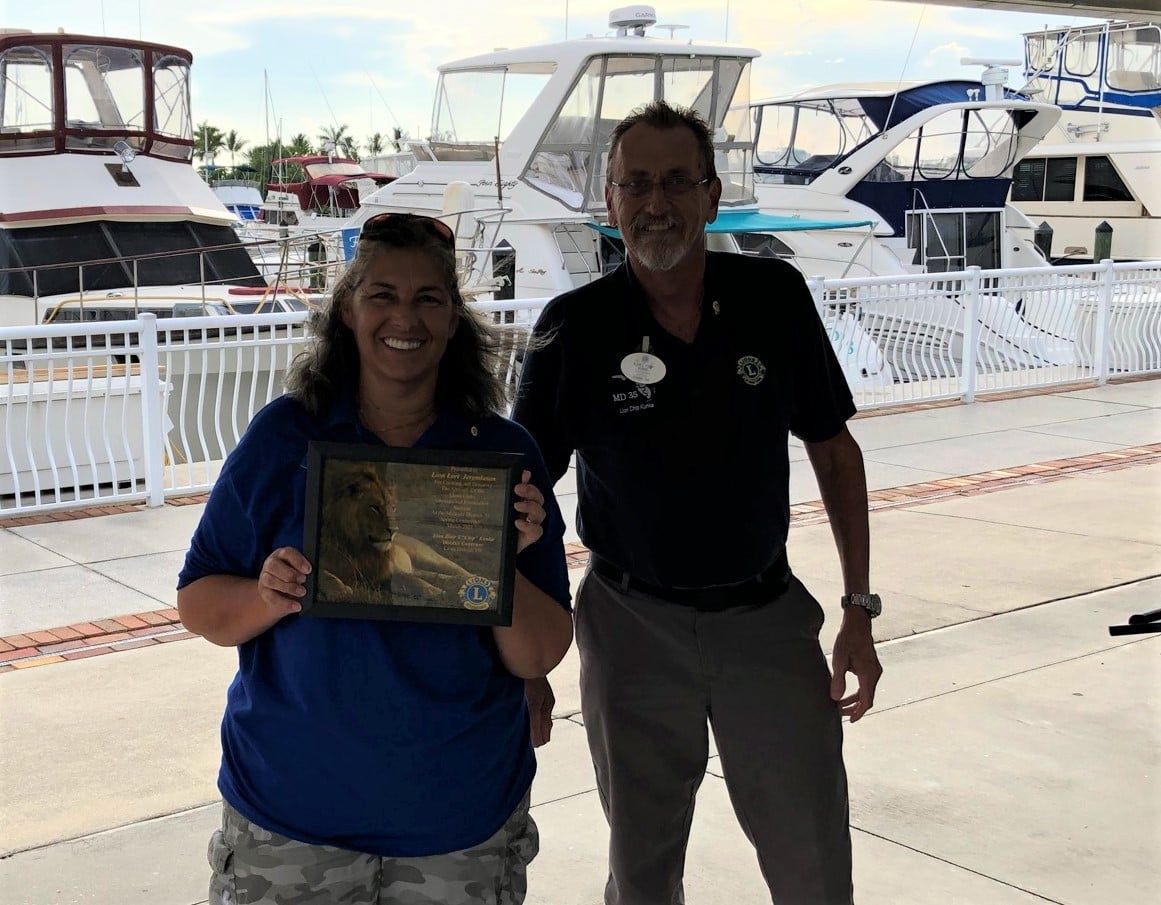 Two people by a marina; woman holds a framed award. Both smile. Boats in the background.