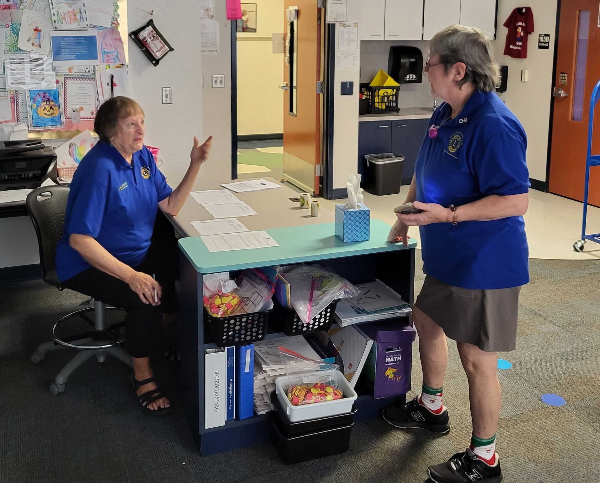 Two women in blue shirts converse at a desk in a brightly lit room. One sits, gesturing.