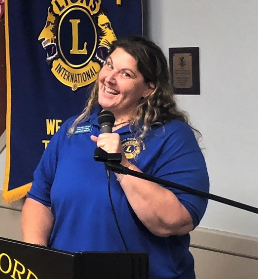 Woman in blue Lions Club shirt speaks into microphone at a podium with a banner.