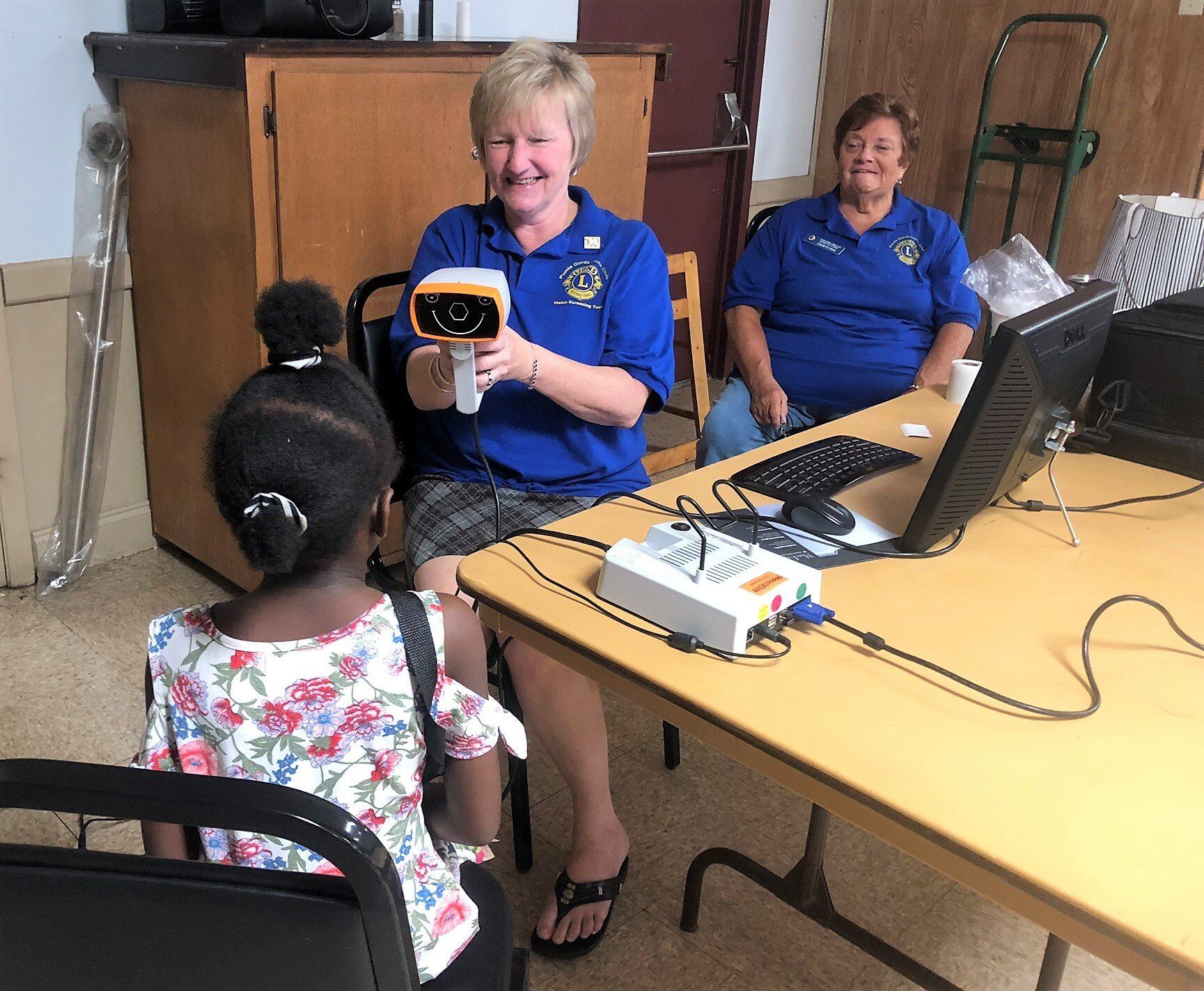 Woman using eye screening device on a child; another woman watches. All are indoors at a table.