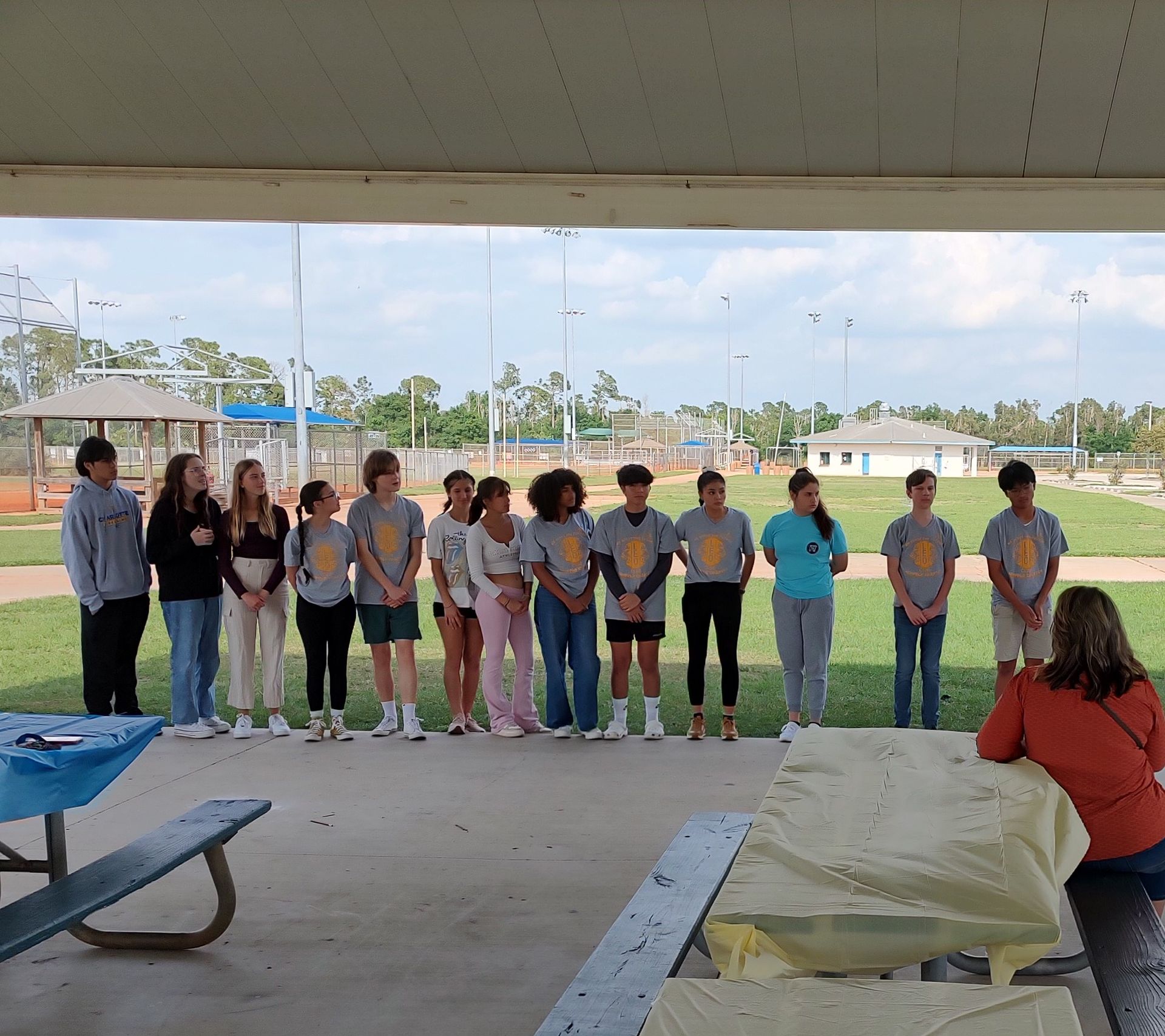 Group of teens in matching shirts stand outdoors with a woman at a picnic table.