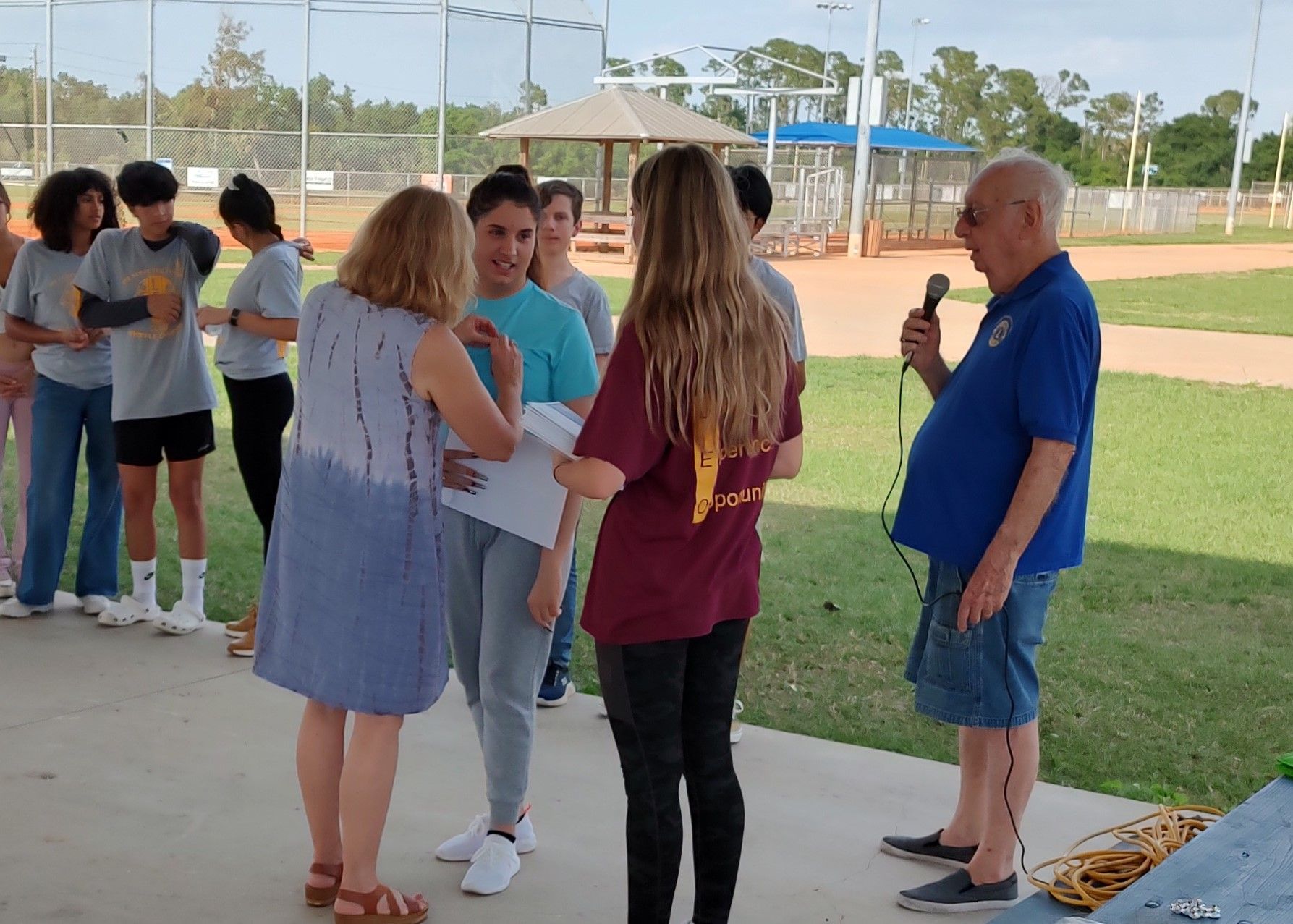 People at an outdoor ceremony: woman handing a paper to a teen, man with a microphone speaks.