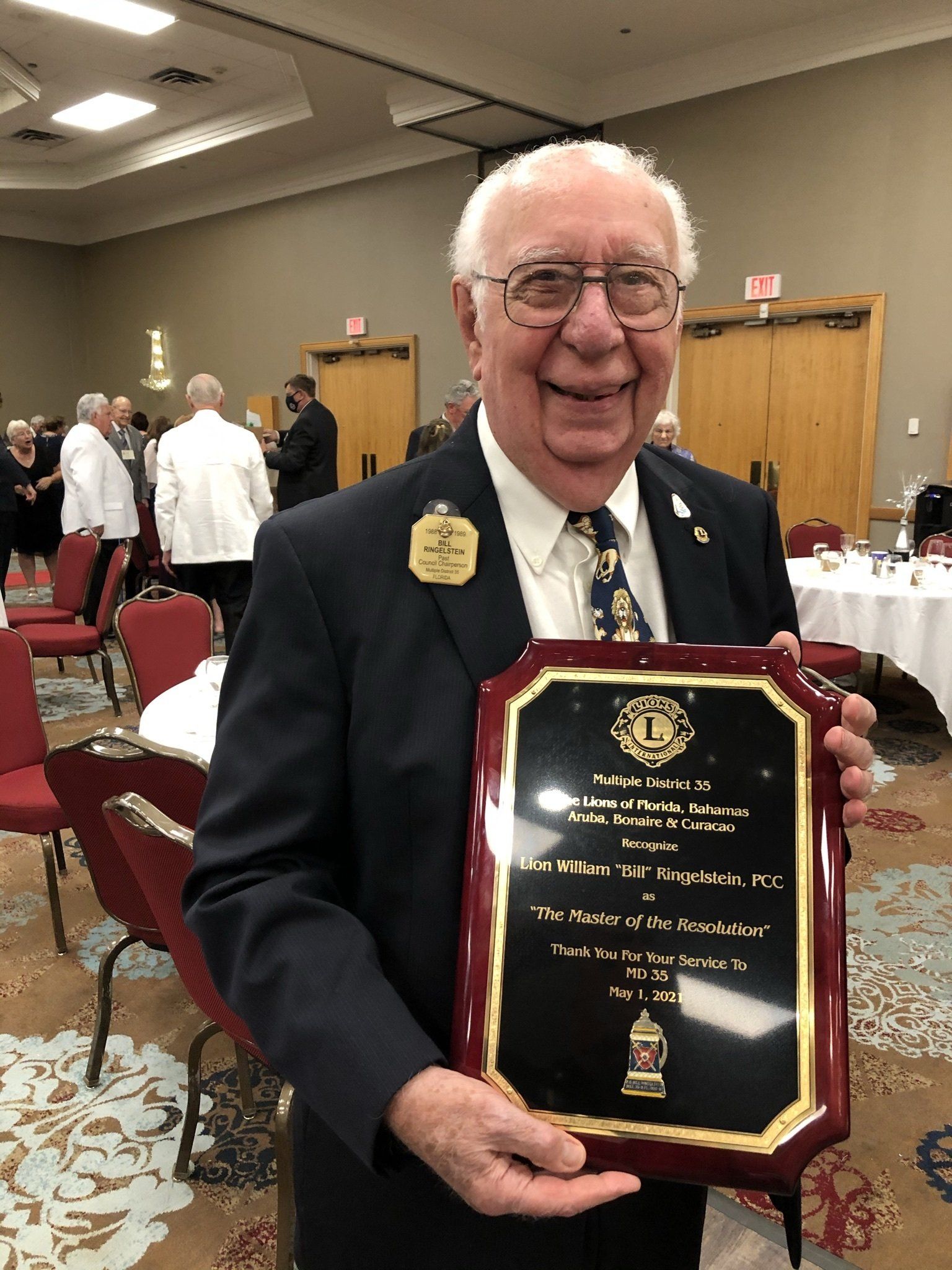Smiling senior man holds plaque at a banquet. He wears a suit and a Lions Club pin.