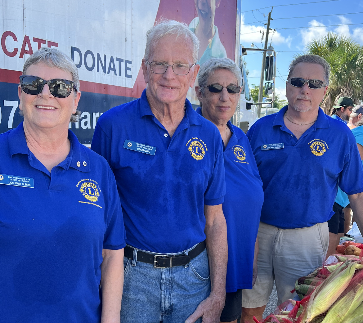 Four Lions Club members in blue shirts standing behind a table of produce, outdoors.