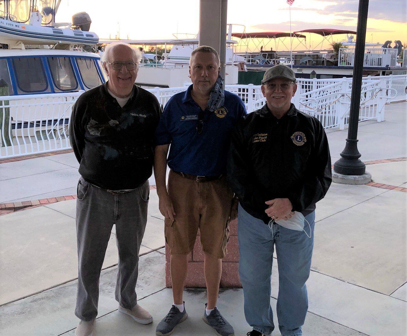 Three men stand outside near a boat. One wears a black sweater, one a blue shirt and shorts, and one a jacket.