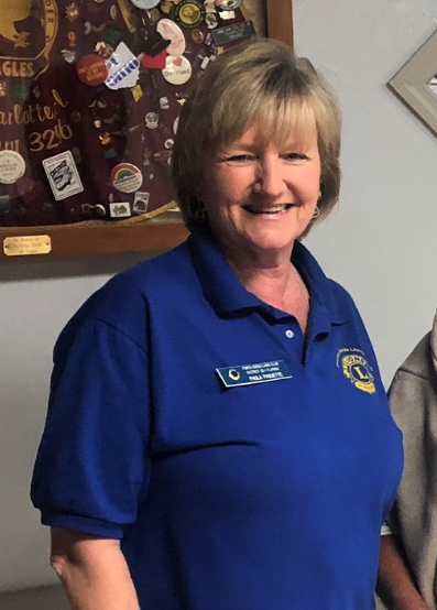 Woman in blue shirt with Lions Club logo, smiling, standing indoors.