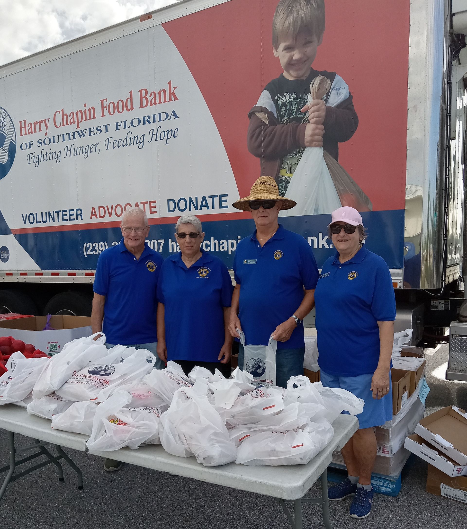 Four people in blue shirts stand behind a table with bags of food; Barry Chaplin Food Bank truck in background.