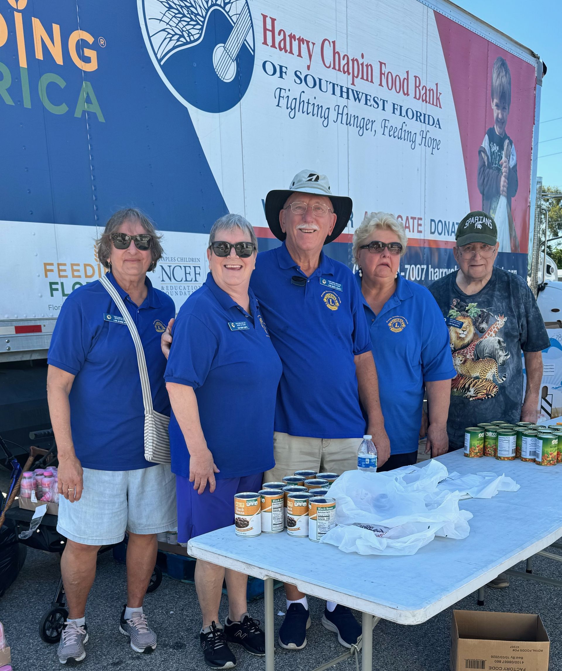 Group of people in blue shirts, standing by table with canned goods, in front of a truck, supporting food drive.
