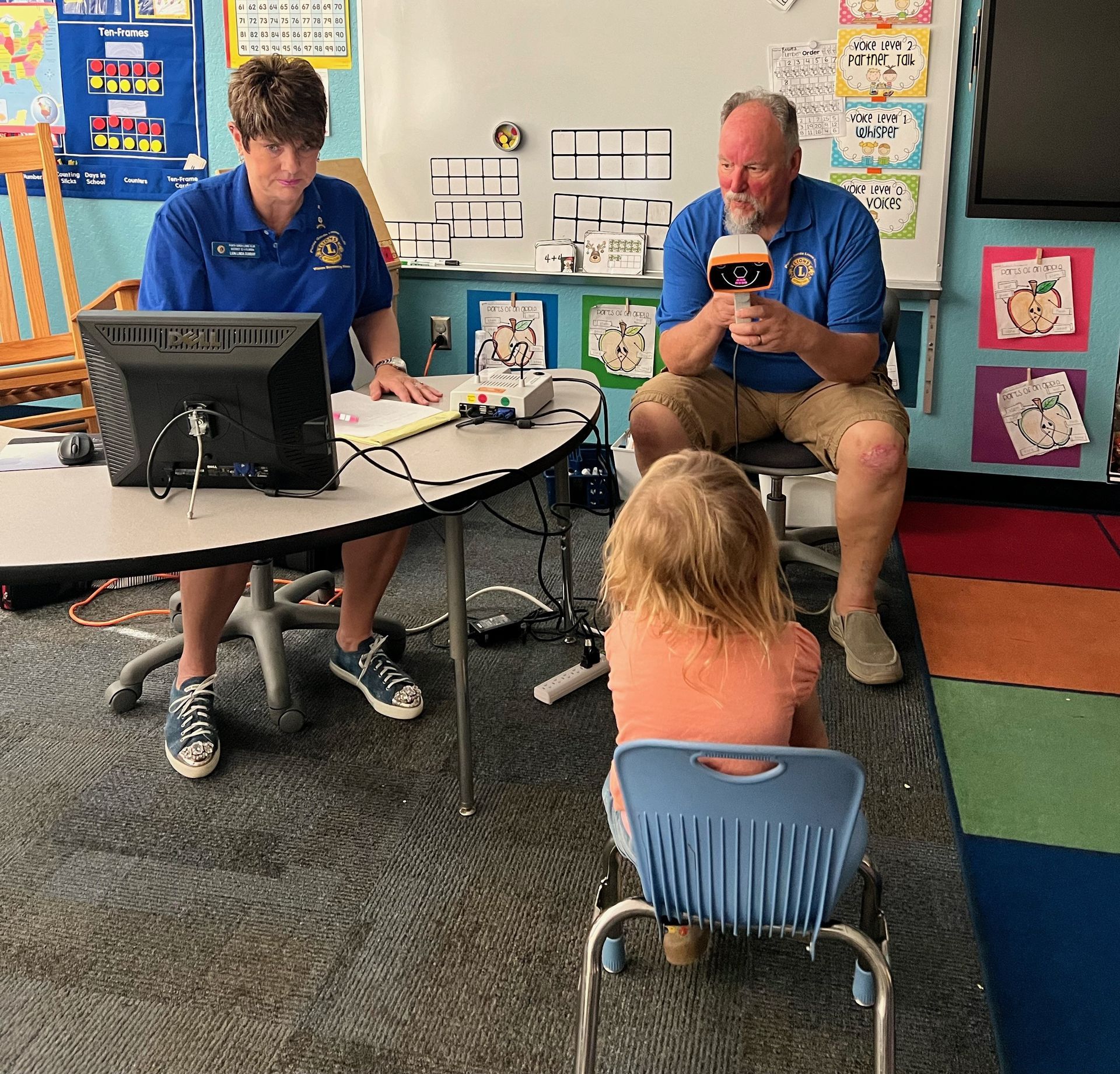 Two adults in blue shirts interview a child in a classroom. Man holds microphone.