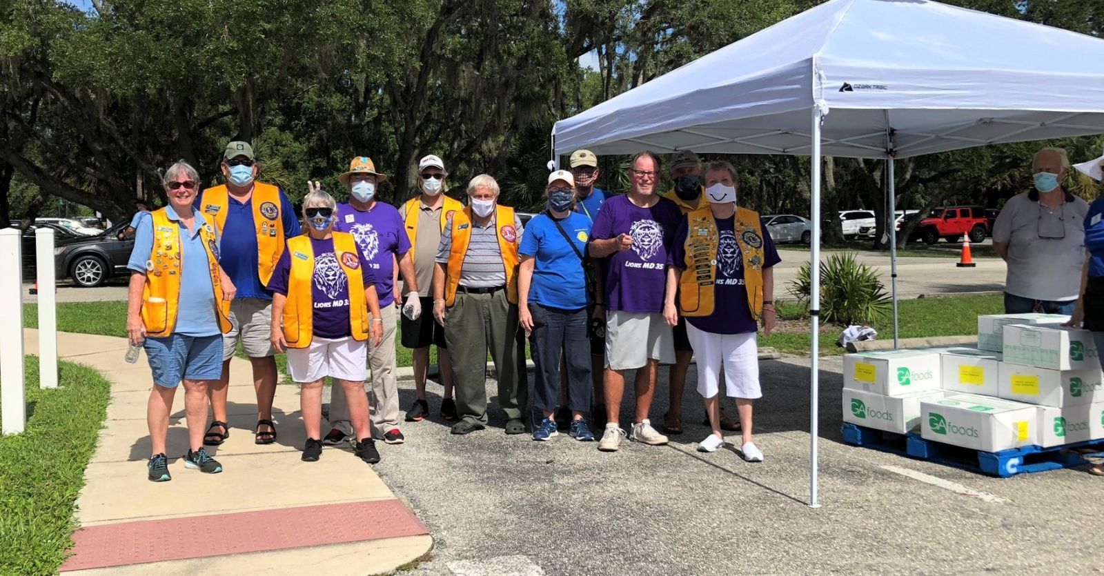 Group of Lions Club members distributing goods under a tent; wearing vests, masks, and shirts.