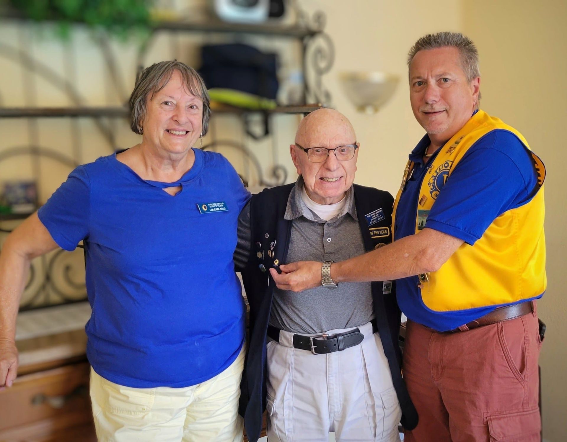 Three people posing: a woman in blue, a man in a suit with pin, and a man in a Lions Club vest; indoors.