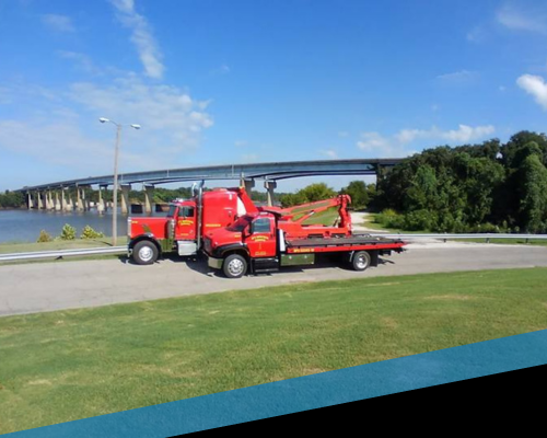 Red tow truck carrying a red tractor on a flatbed near a bridge by the water under a blue sky