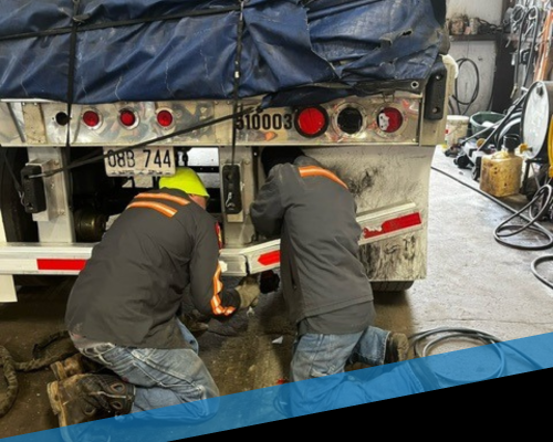 Two workers in safety vests crouch under a truck, repairing its underside in a garage
