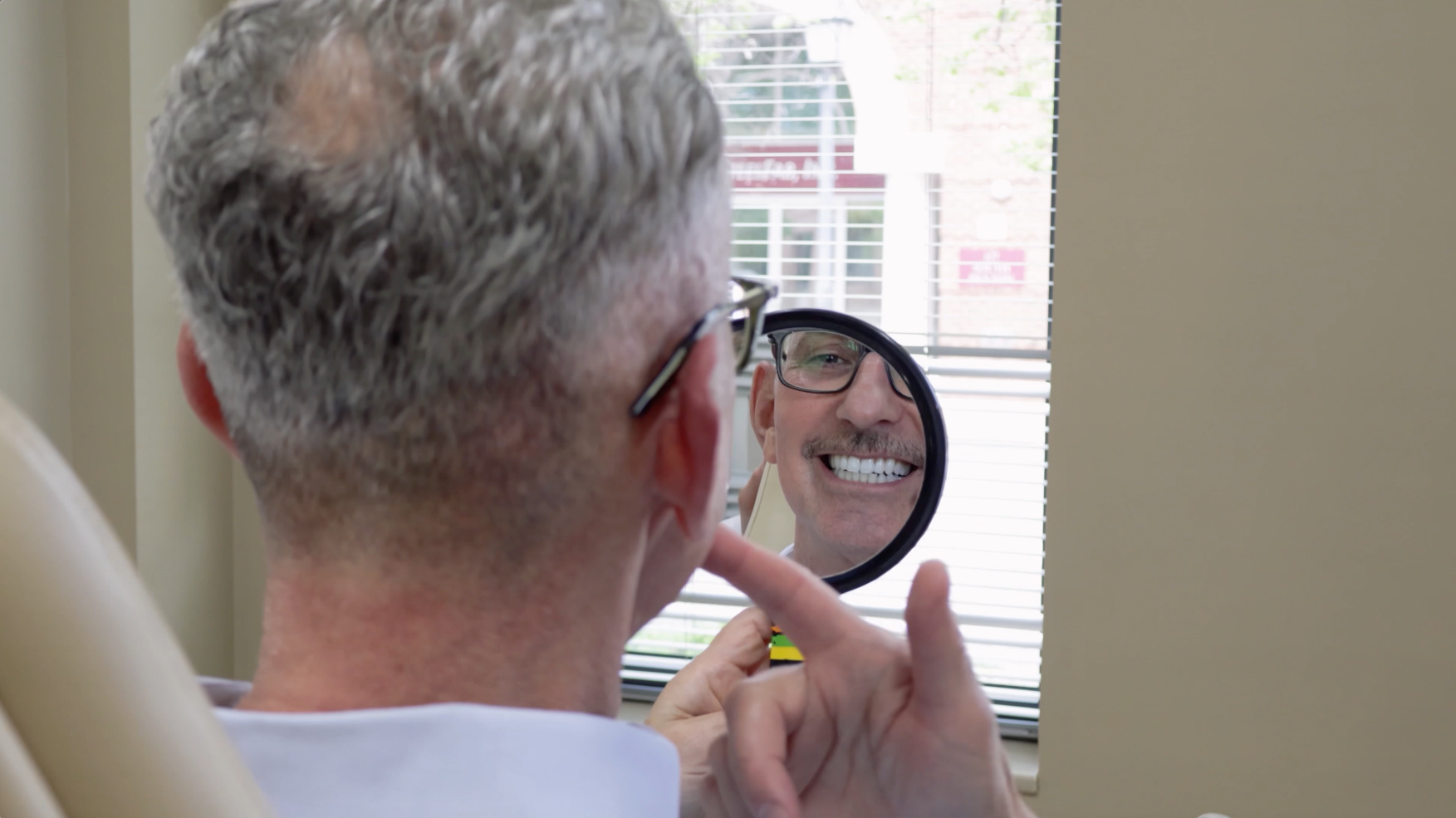 Man smiles, checking his teeth in a mirror at a dentist's office.