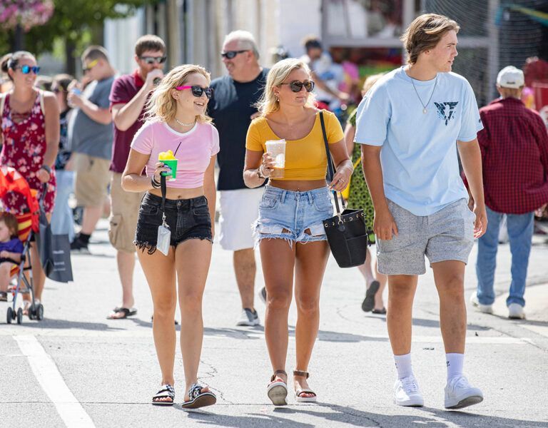Three friends walking outdoors in bright sunlight, two holding drinks and wearing casual summer clothes.