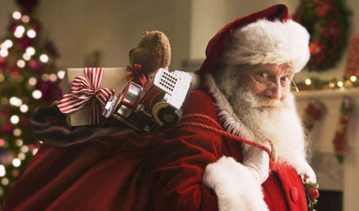 Santa Claus in a red suit carries a large sack of toys and gifts over his shoulder in a decorated room with a Christmas tree.