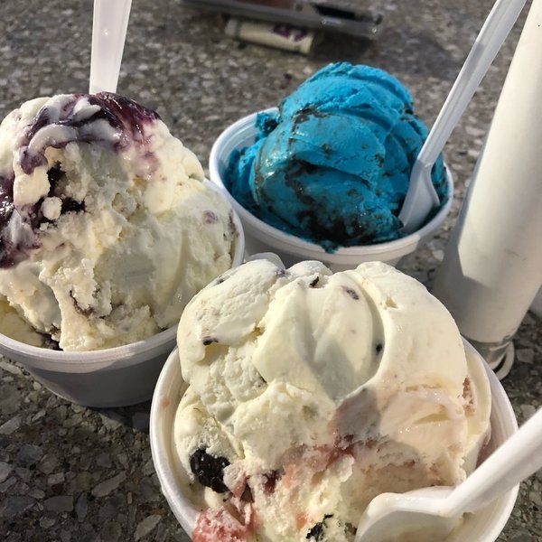 Three cups of colorful ice cream outdoors: blue, white, and mixed berry scoops with spoons.