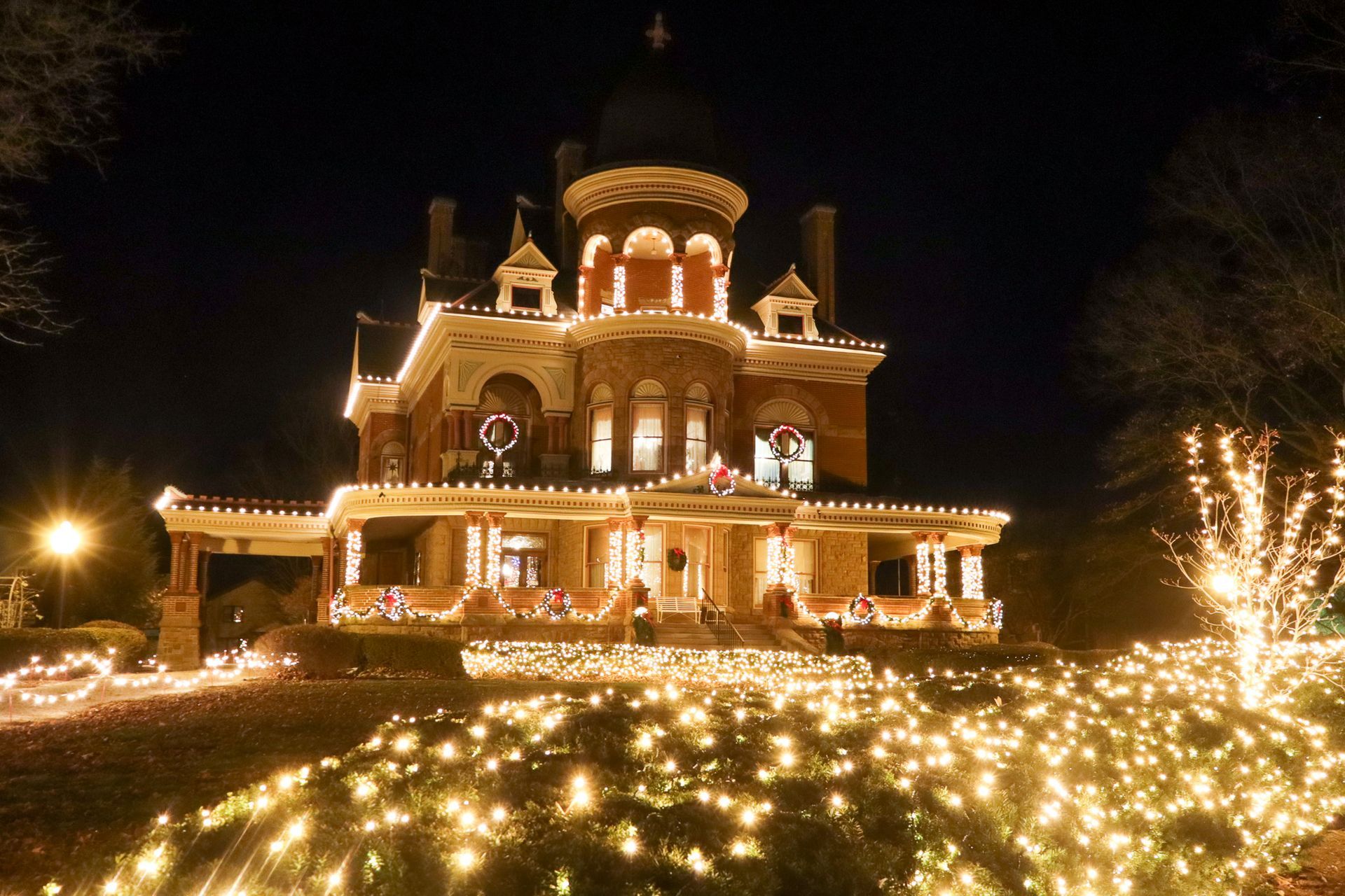 A historic, multi-story brick house illuminated at night with extensive Christmas lights decorating the exterior and lawn.