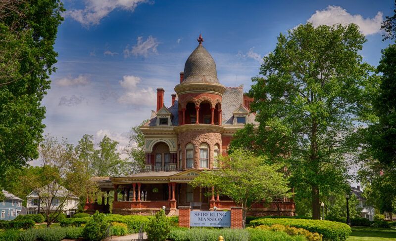 Victorian-style historic house with a domed turret, surrounded by green trees and landscaping under a blue sky