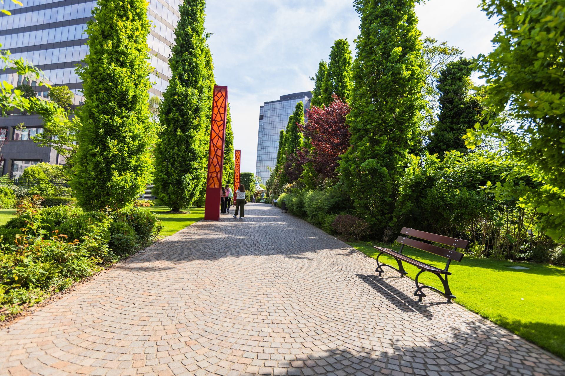 Stone path in a park lined with trees and a bench. Two people walk towards buildings.