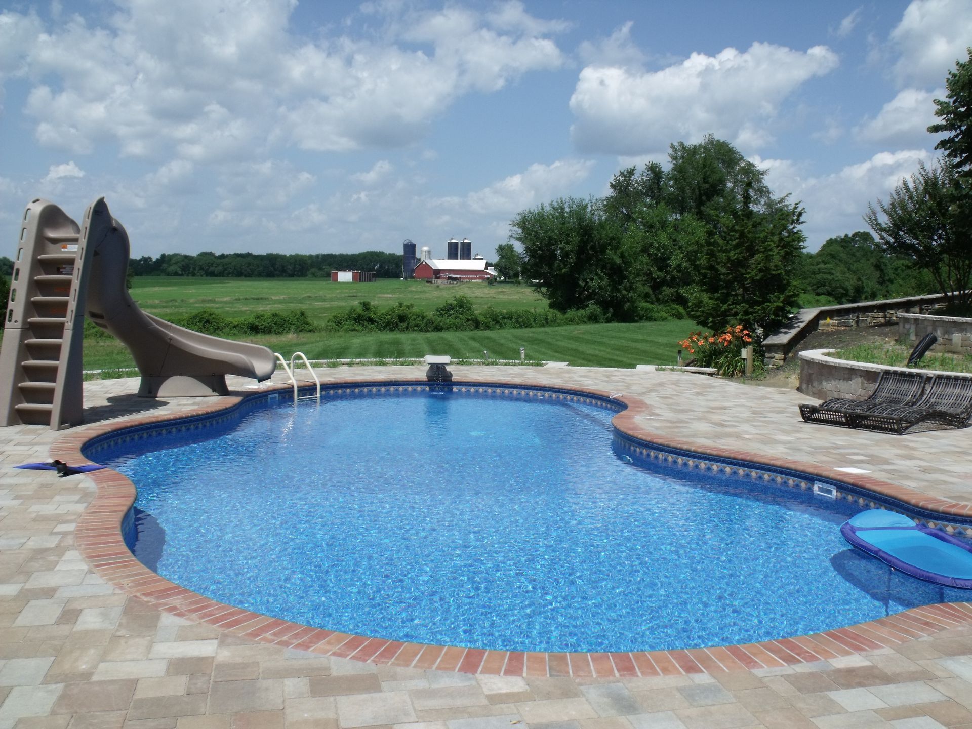 Pool with slide and brick patio in front of a green field with a distant red barn.
