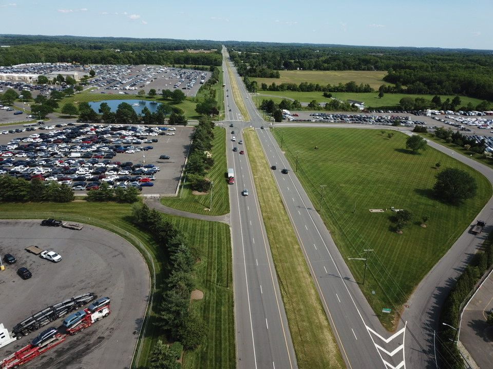 An aerial view of a highway with multiple lanes, surrounded by parking lots and greenery under a blue sky.