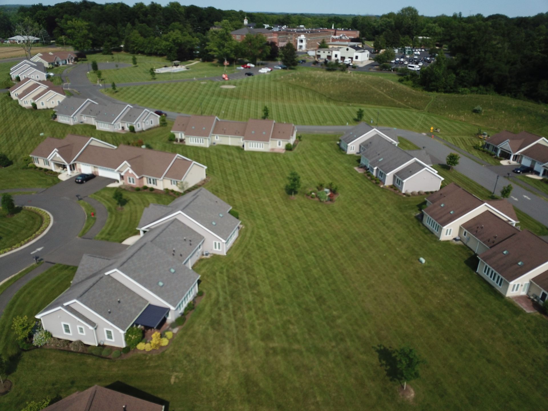 Aerial view of a suburban neighborhood with rows of houses, green lawns, and a larger building in the background.