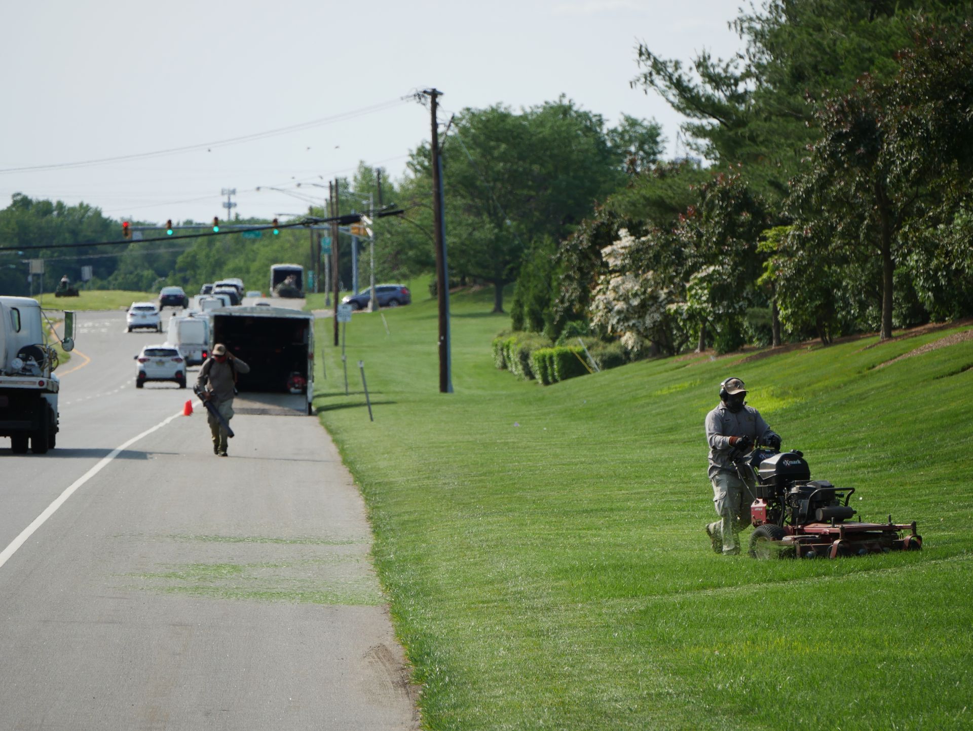Lawn care worker mowing a grassy slope next to a road. Another person walks by a trailer.