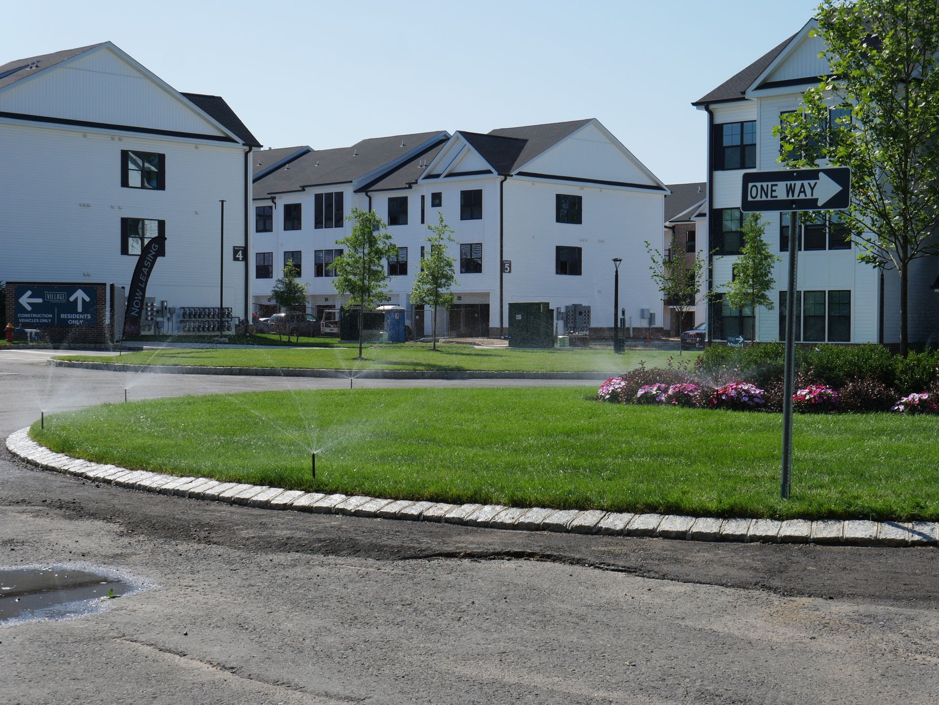 Buildings surround a circular lawn with sprinklers, a one-way street sign, and a paved road.