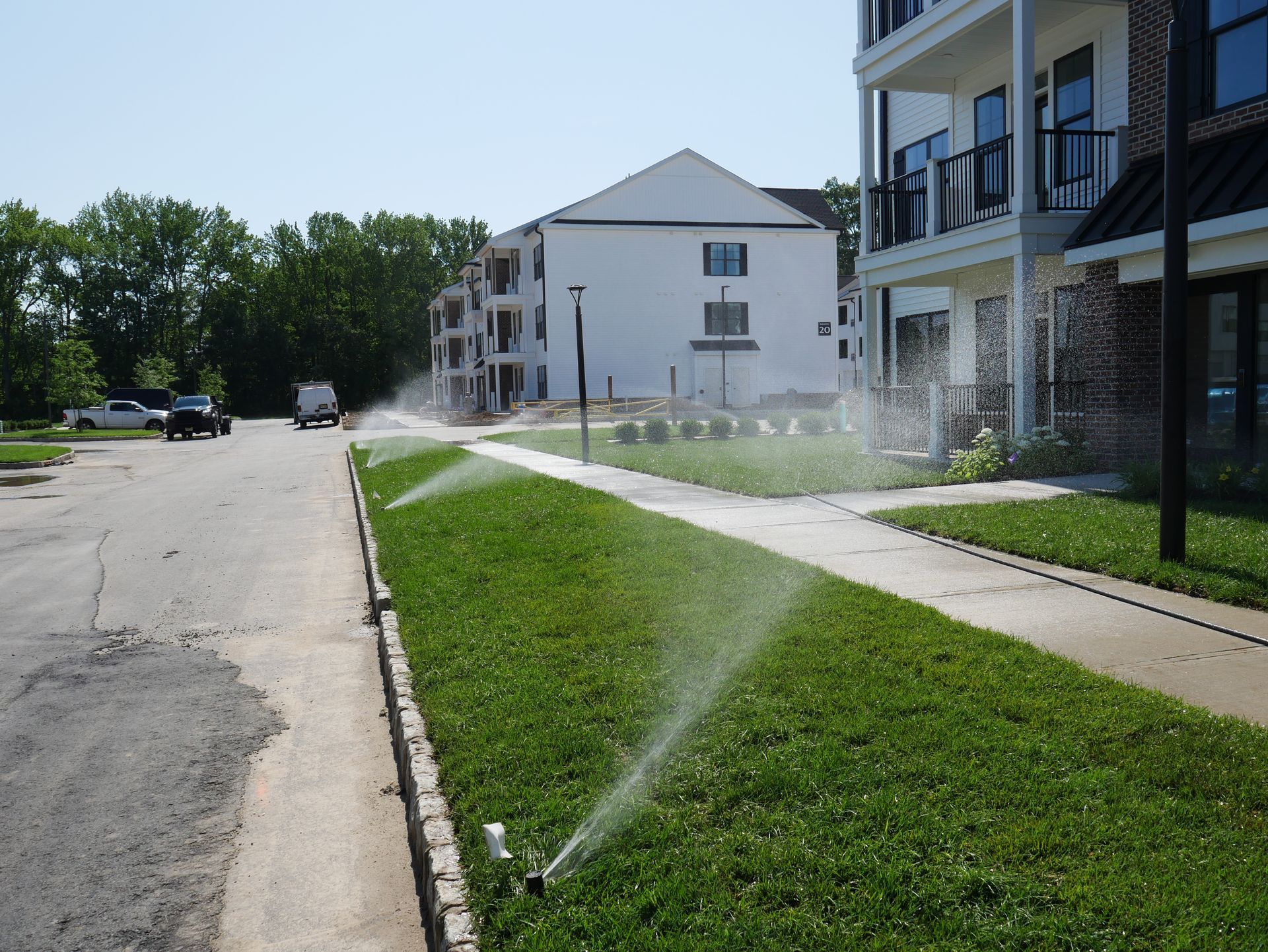 Sprinklers watering green grass along a road, in front of white and dark buildings on a sunny day.