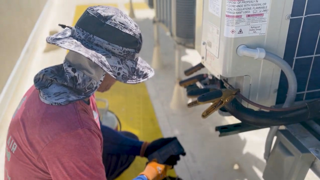 Person wearing a sun hat and gloves works on wall-mounted equipment outdoors.