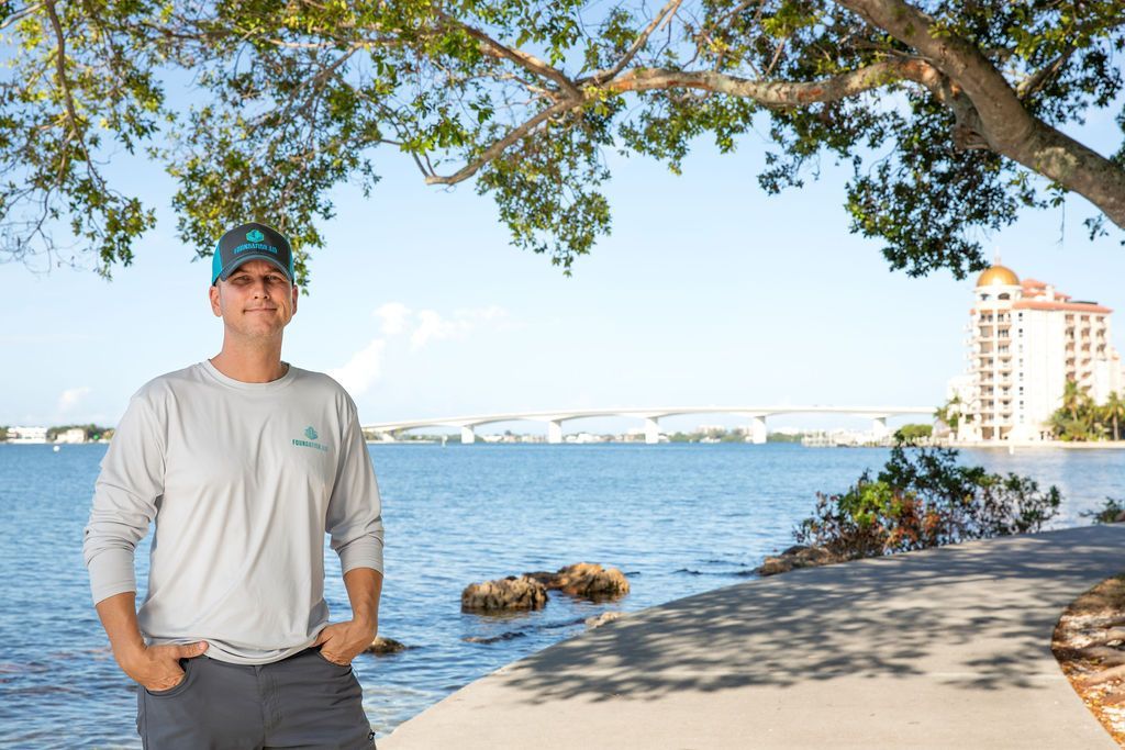 Man standing on a waterfront path beside blue water, trees overhead, and city buildings in the distance.