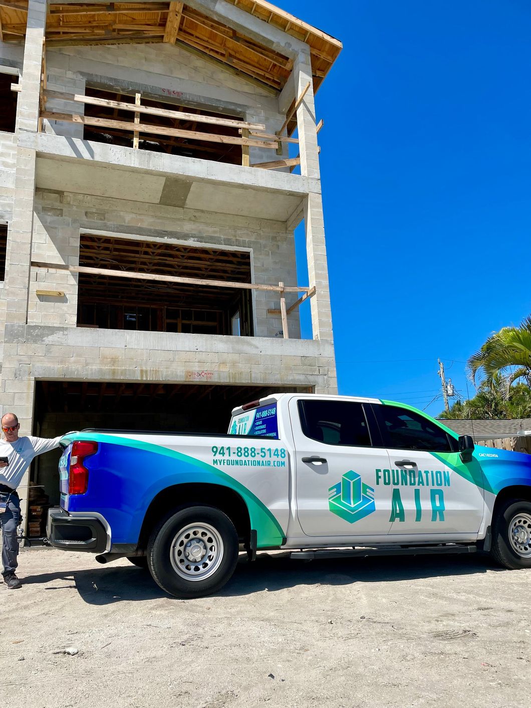 White and teal pickup truck parked in front of an unfinished concrete building under a bright blue sky