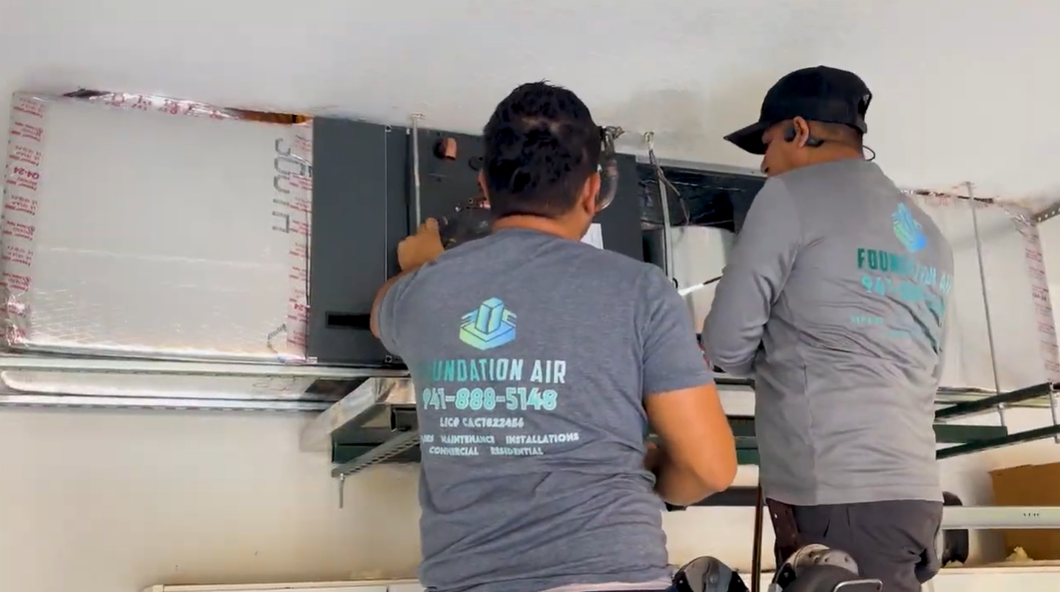 Two technicians in gray shirts work on a wall-mounted air-conditioning unit indoors.