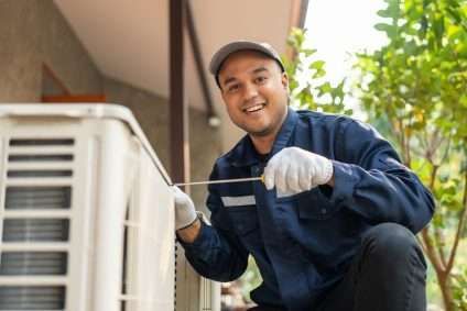 Technician kneeling beside an outdoor AC unit, wearing gloves and a cap while smiling and inspecting it.