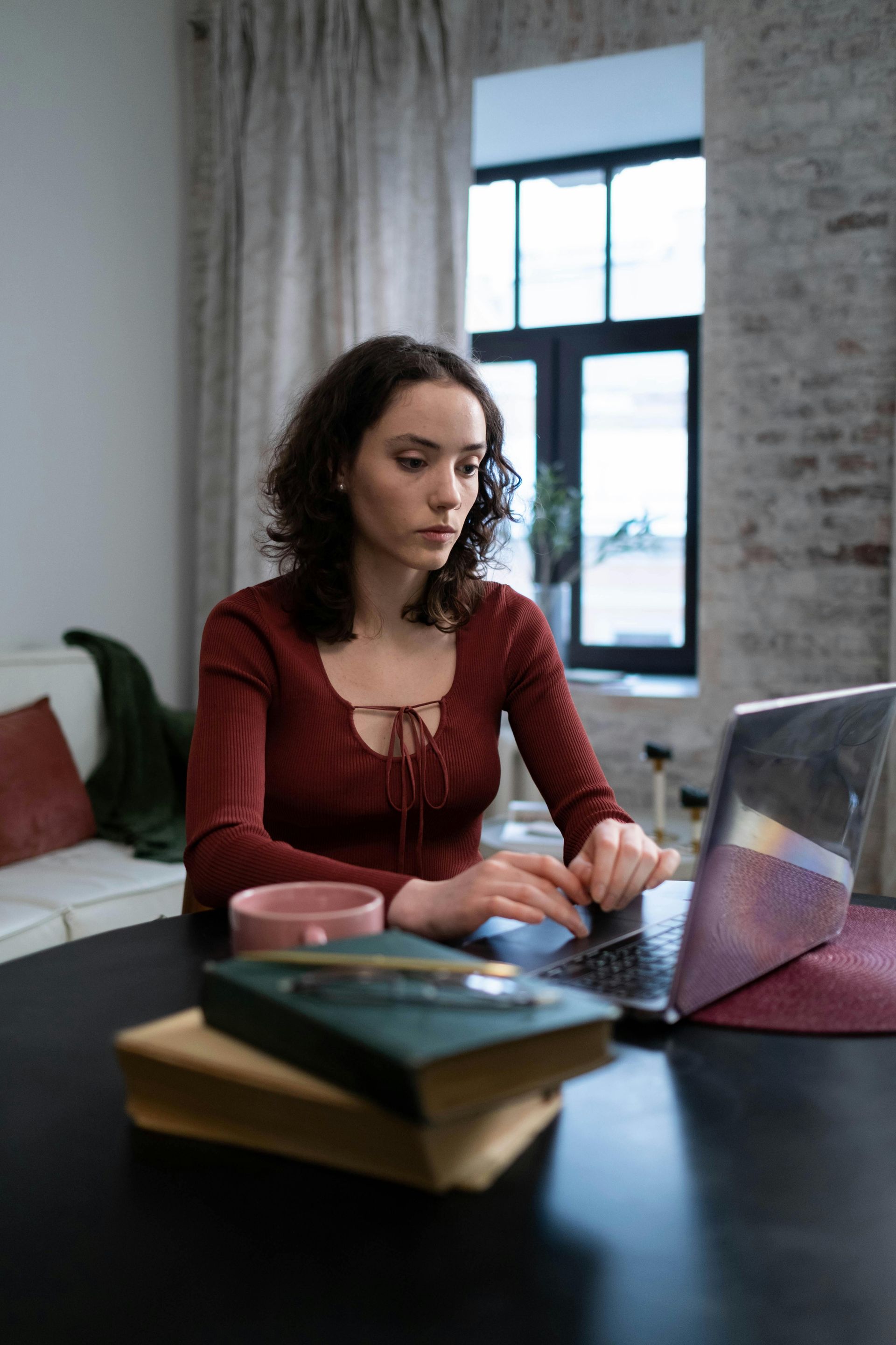 Woman working on a laptop at a table in a bright room with books and a mug.
