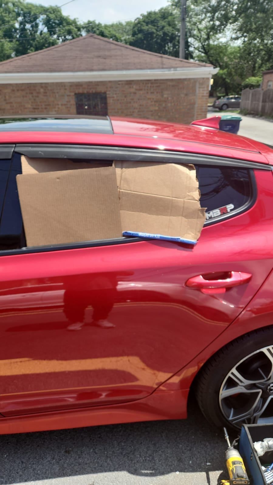 Red car with cardboard blocking a rear window, parked outdoors.