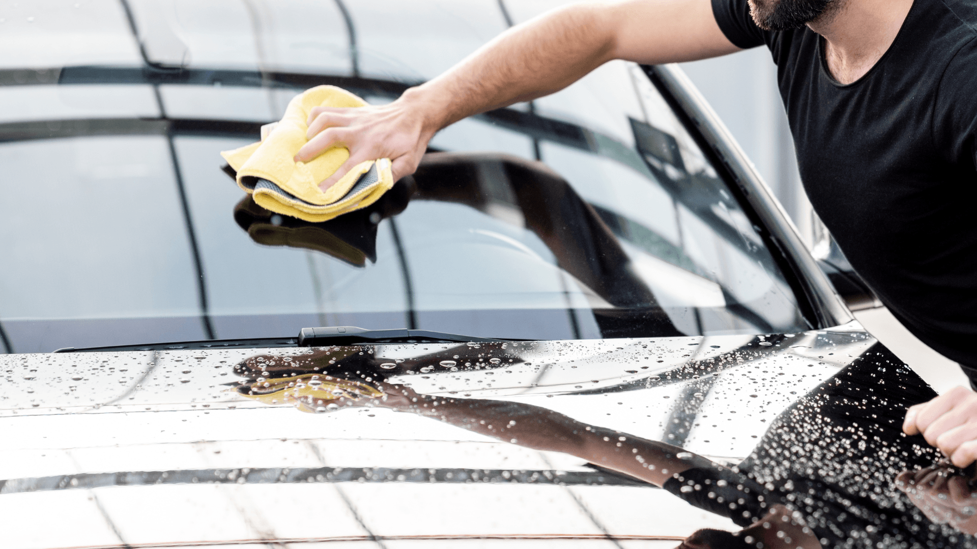 Man wiping the windshield of a black car with a yellow cloth, outdoors.