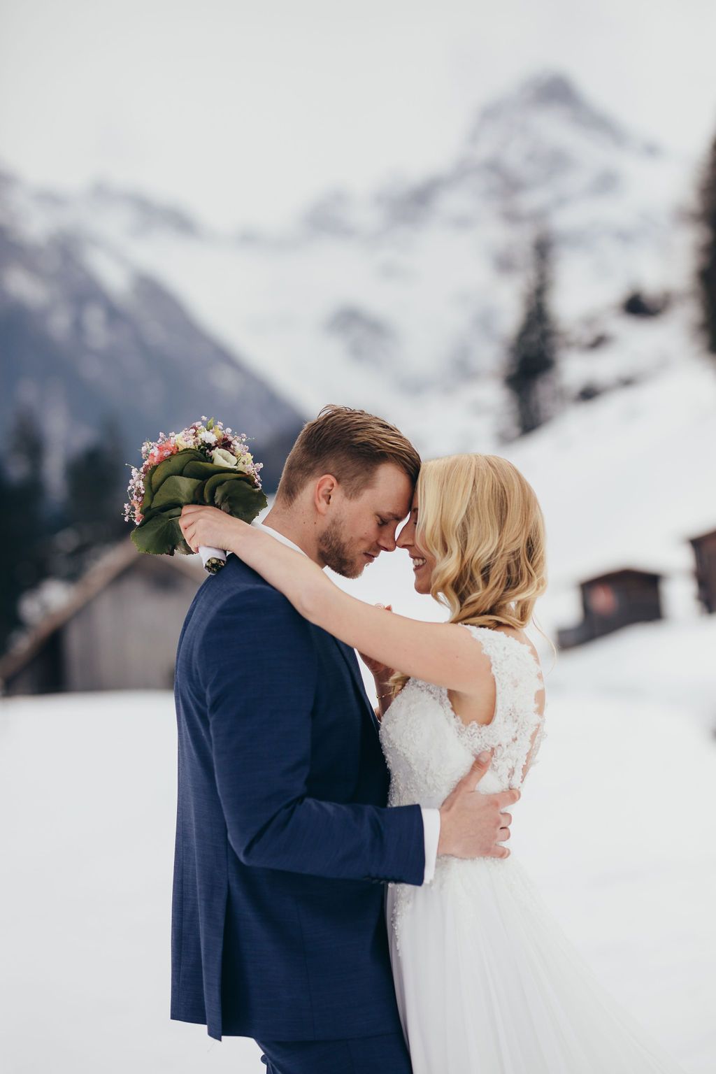 Eine Braut und ein Bräutigam küssen sich im Schnee mit Bergen im Hintergrund.