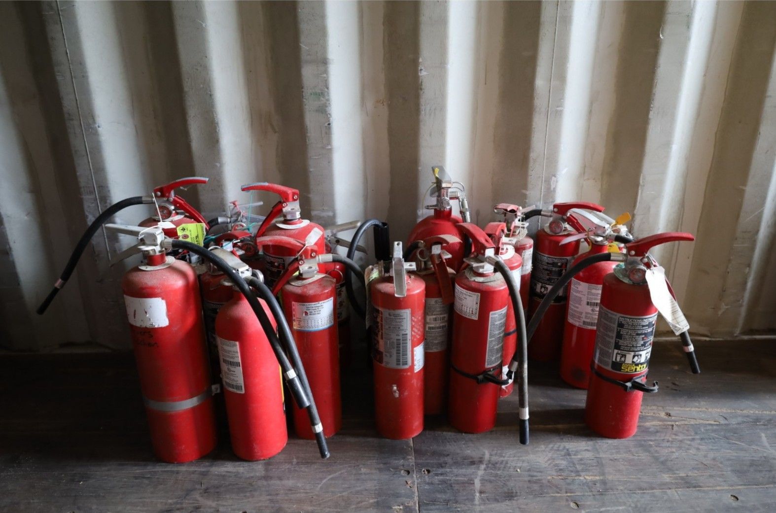 A collection of red fire extinguishers with black hoses, arranged in a row against a corrugated metal wall.