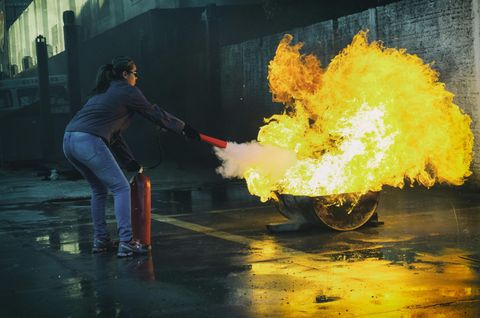 Woman using fire extinguisher on a large fire, outdoor setting.
