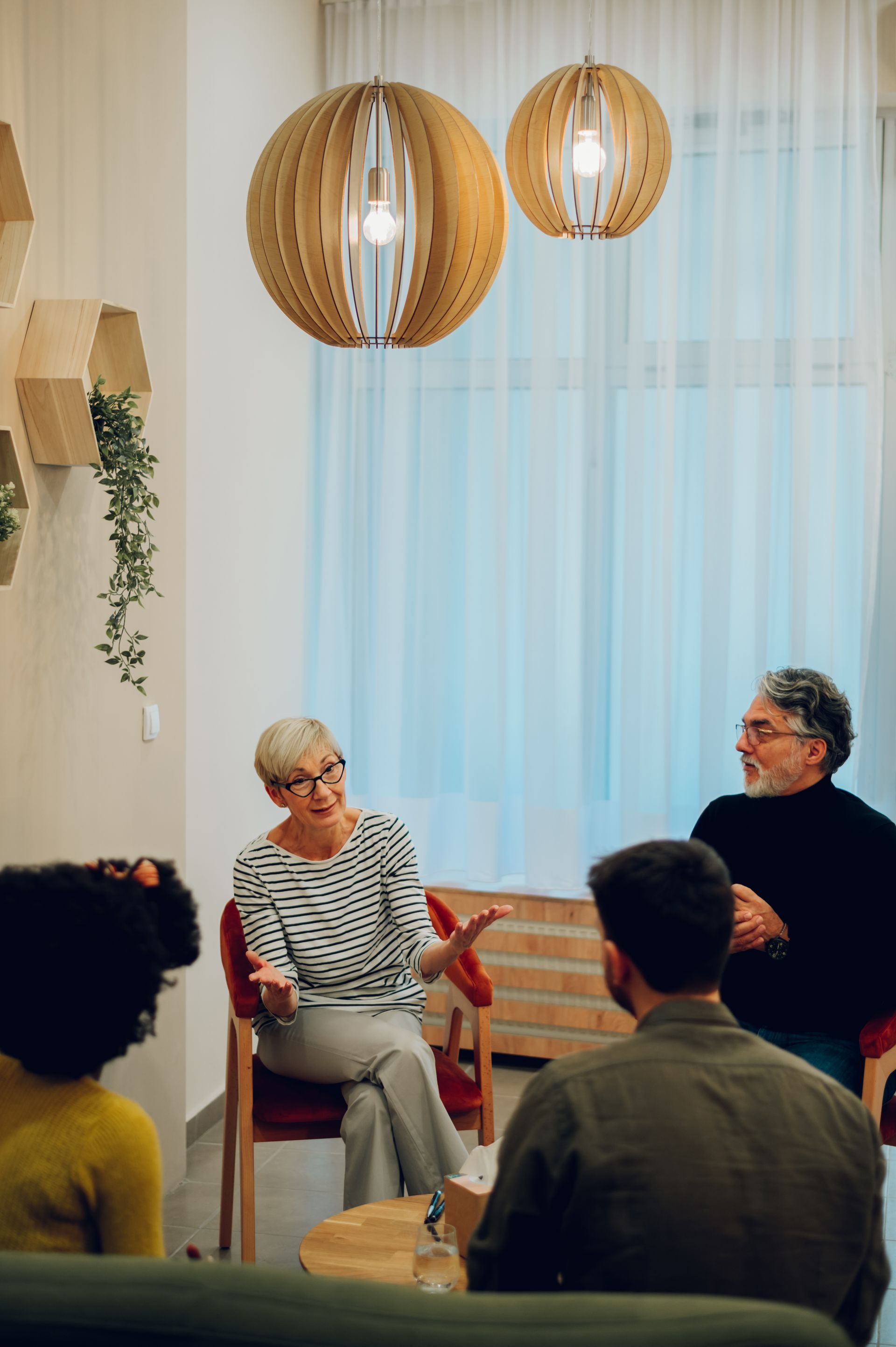 Two people speaking in a cozy room with hanging pendant lights and soft curtains