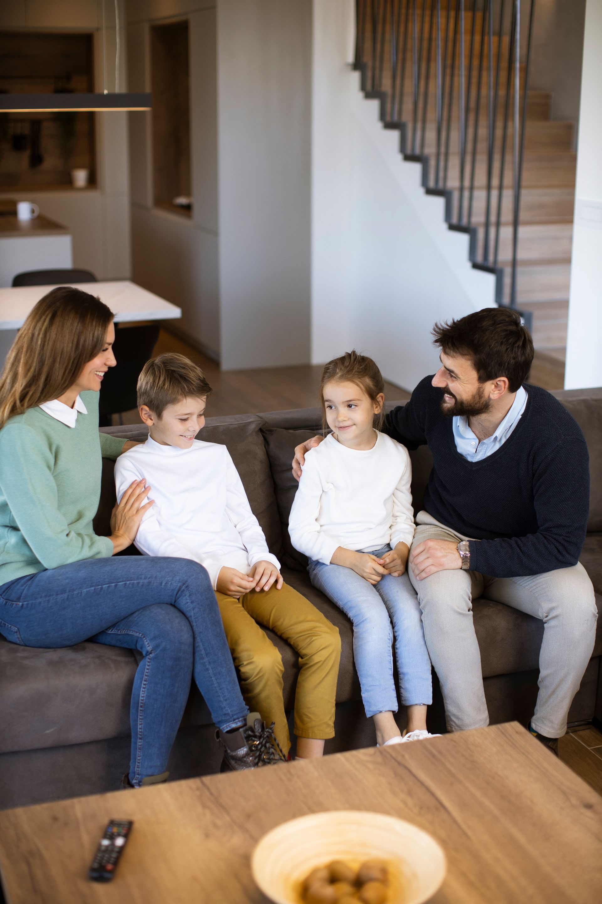 Family sitting together on a couch in a living room, chatting near a staircase.