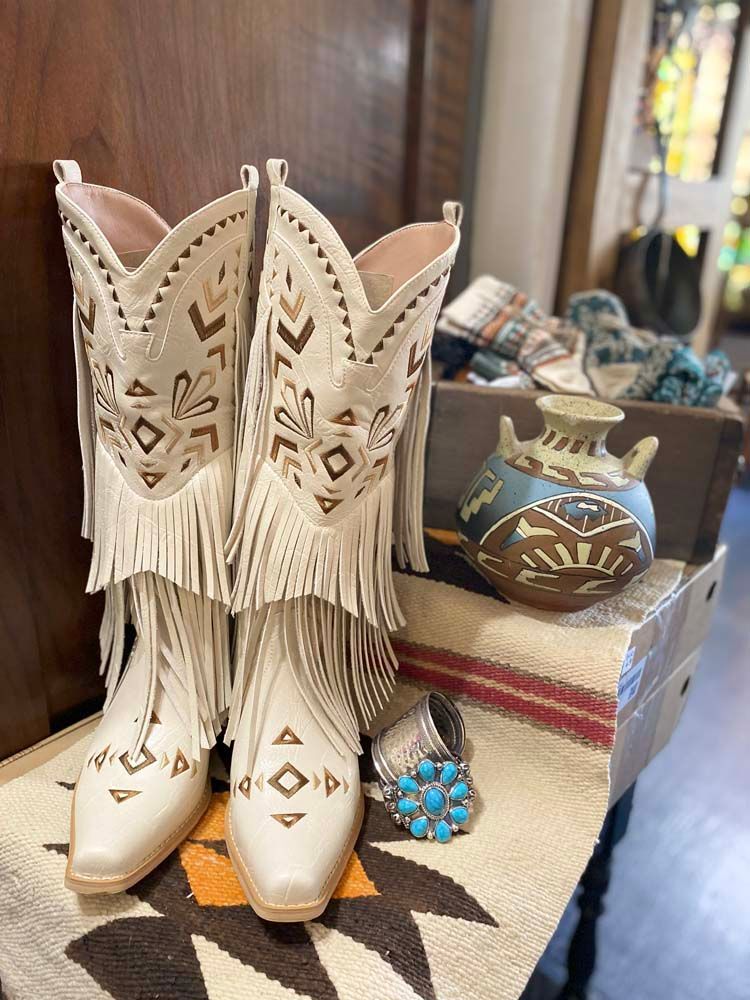 A pair of white cowboy boots with fringe are sitting on a table.