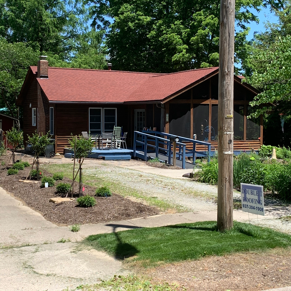 Small brown house with red roof, blue ramp, screened porch. Gravel driveway, lush green yard, and wooden pole.