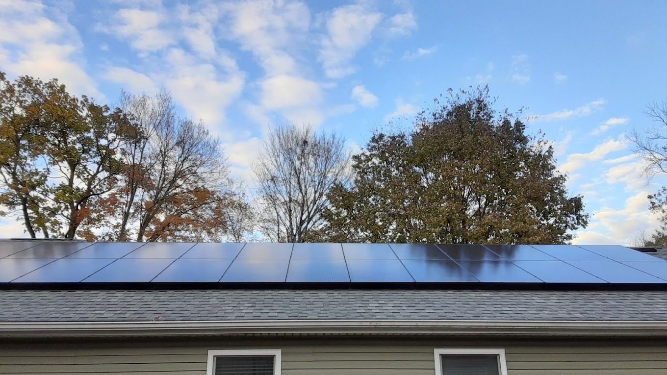 Solar panels on a house roof with trees and blue sky with clouds in the background.