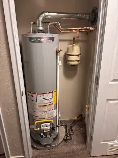 A gray gas water heater sits in a utility closet with a copper piping system and a small expansion tank mounted above.