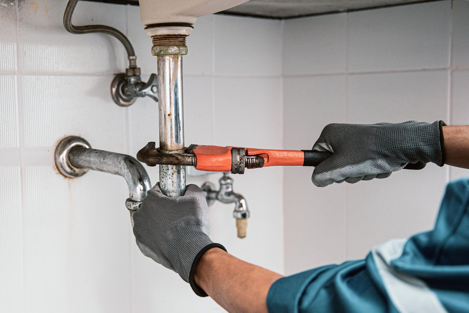 A person wearing gray work gloves uses an orange pipe wrench to tighten a metal drainpipe under a bathroom sink.