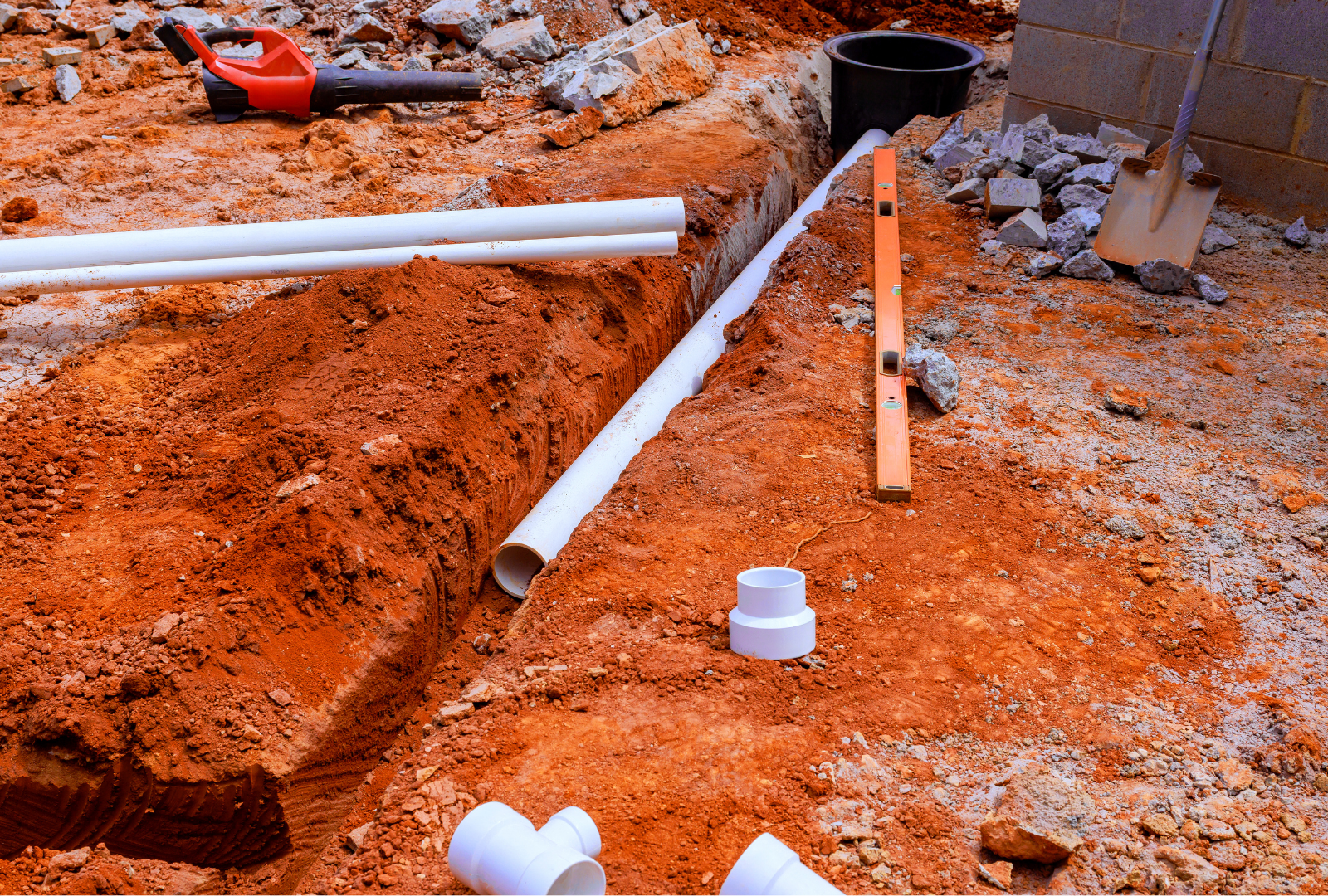 A construction site showing white PVC pipes, a level, and plumbing fittings placed in a trench with orange soil.